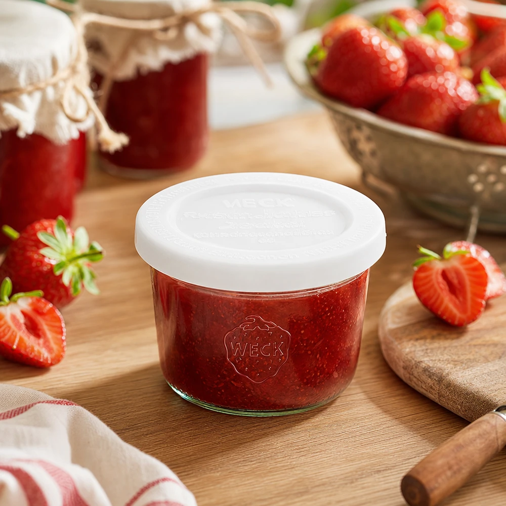 WECK 80ml bekers RR60 gevuld met aardbeienjam op een houten plank omringd door verse aardbeien en jampotten